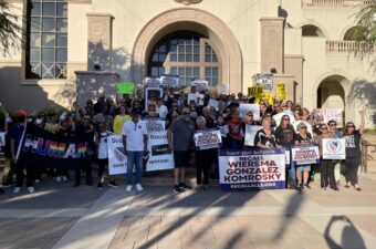 A large group of people pose for a photograph in front of a school holding up protest signs.